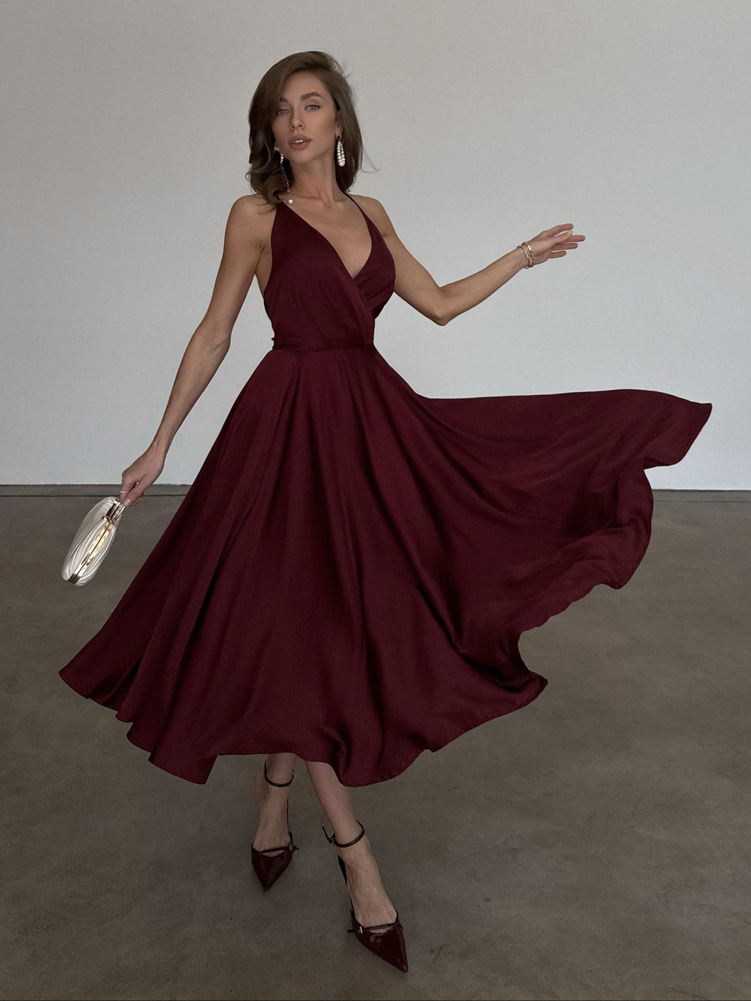 A woman wearing a flowing deep burgundy midi dress with a V-neckline and halter-style straps, holding a small metallic clutch in a minimalist studio setting.