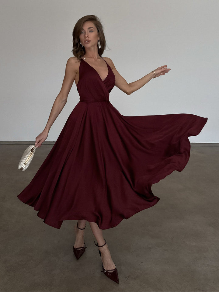 A woman wearing a flowing deep burgundy midi dress with a V-neckline and halter-style straps, holding a small metallic clutch in a minimalist studio setting.