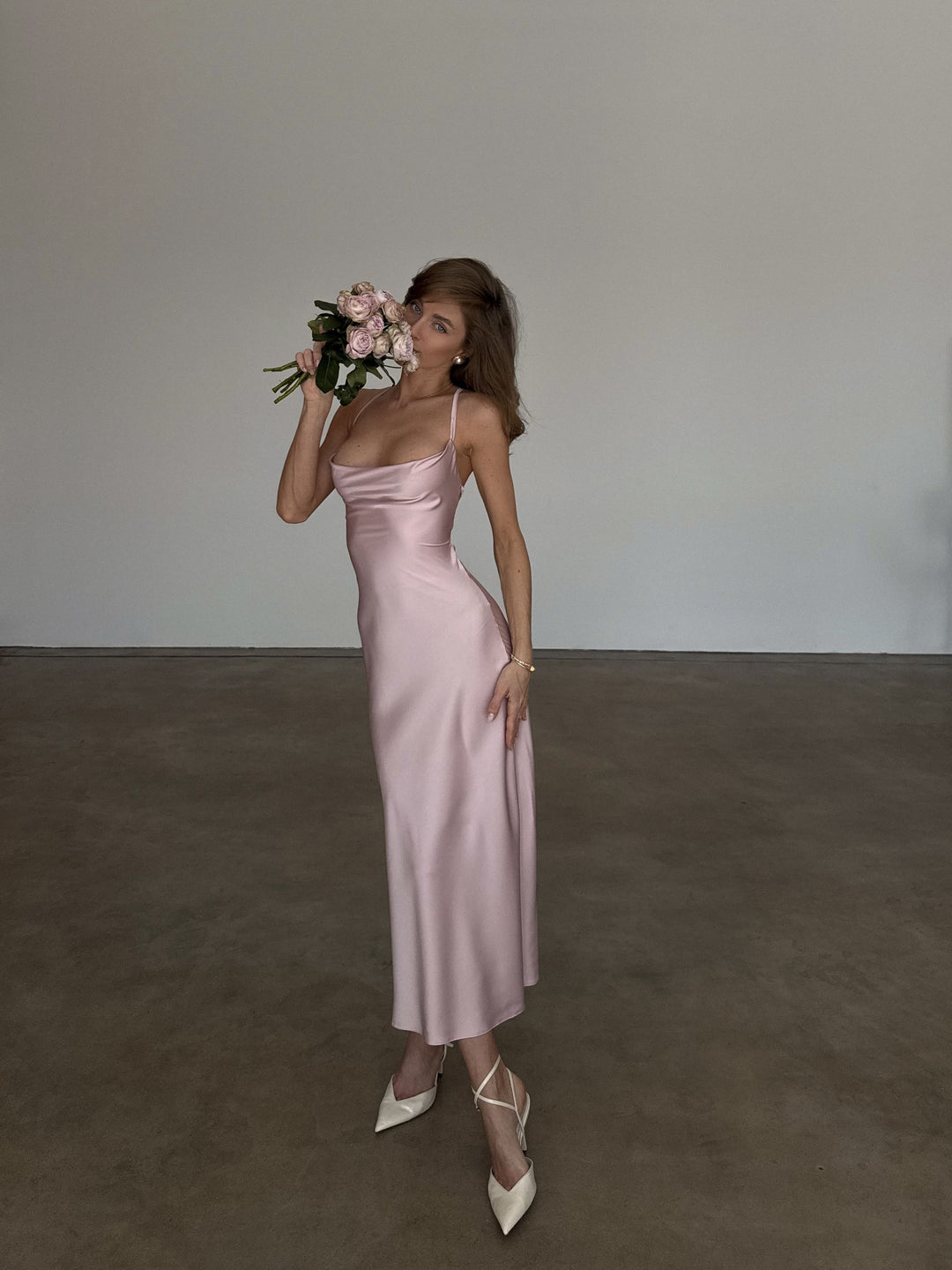 Dusty pink silk satin slip dress with draped cowl neckline and spaghetti straps, full-length front view, bias-cut midi skirt worn with ivory pointed heels and a bouquet of pink garden roses.