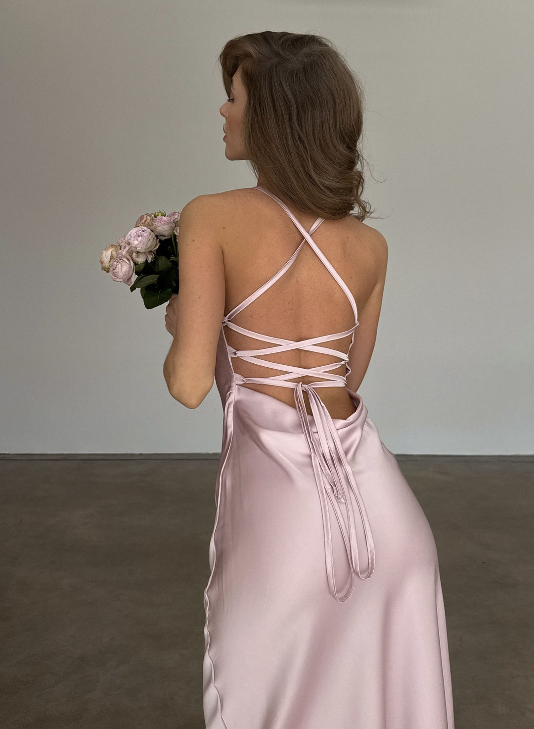Dusty pink silk satin slip dress, criss-cross back straps and lace-up open back detail, satin ties falling over the bias-cut skirt, model glancing over her shoulder.