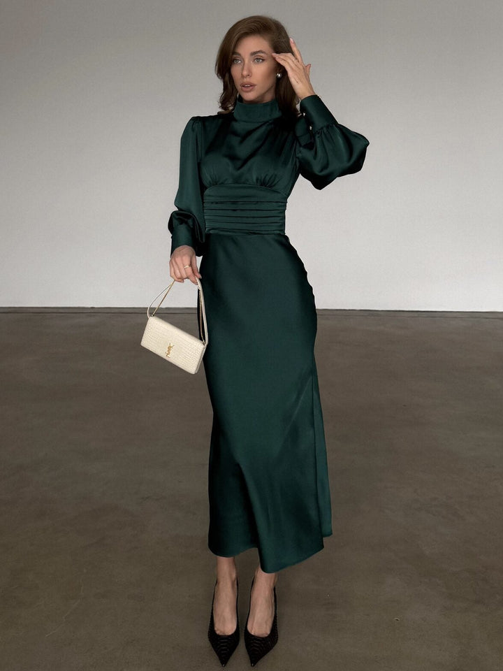 A woman stands in a minimalist studio wearing a dark green satin midi dress with a high neck, puffed long sleeves, and a gathered waist. She is holding a small cream-colored handbag and wearing black pointed-toe heels.