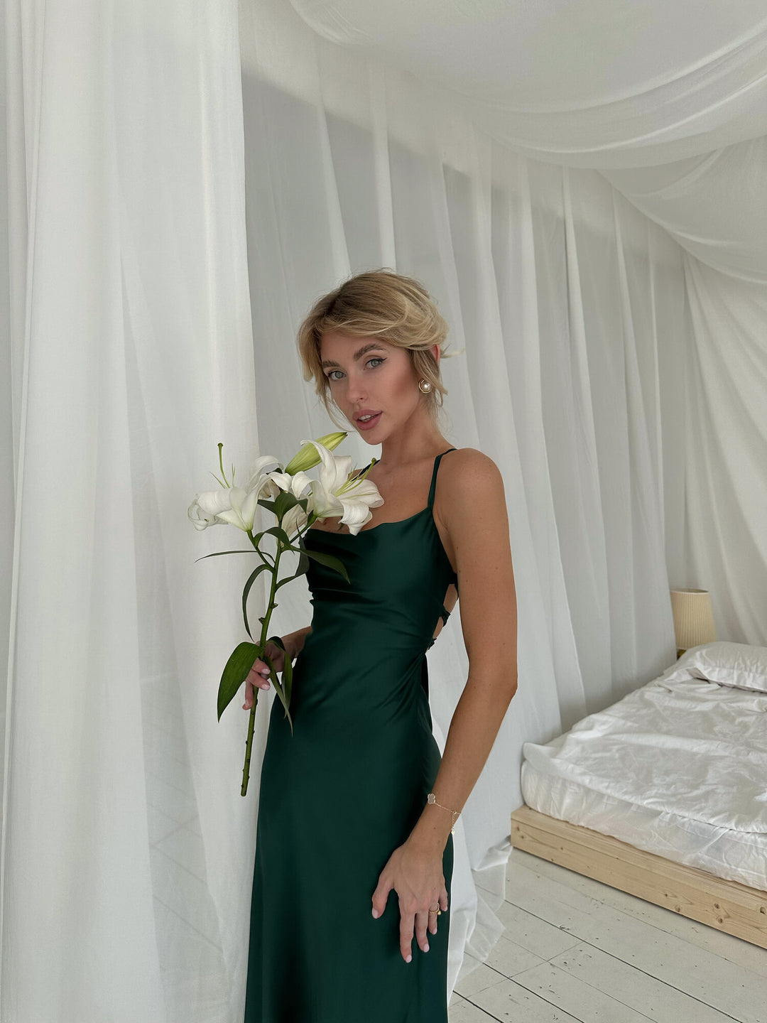 Woman in a green dress holding white flowers in a room with white curtains and a bed.