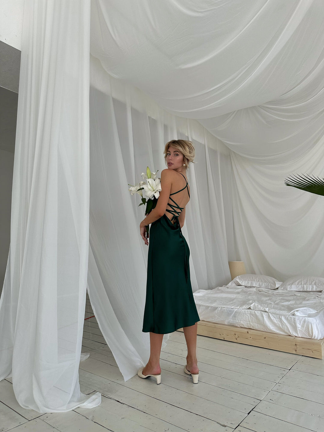 Three-quarter front view of emerald green satin slip dress with cowl draped neckline and spaghetti straps. Bias-cut midi