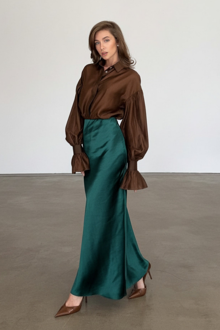 Full-length front view of a model in a studio wearing a high-waisted forest green satin midi skirt paired with a brown sleeved blouse.
