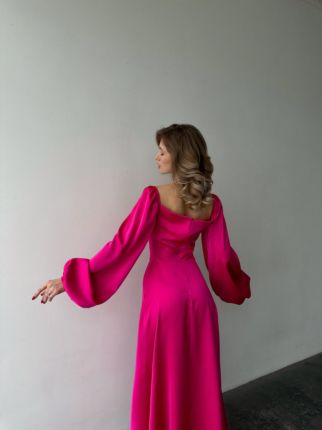 Woman modeling a bold hot pink silk maxi gown with voluminous sleeves and a high slit. A luxurious look for a sophisticated evening reception or party.