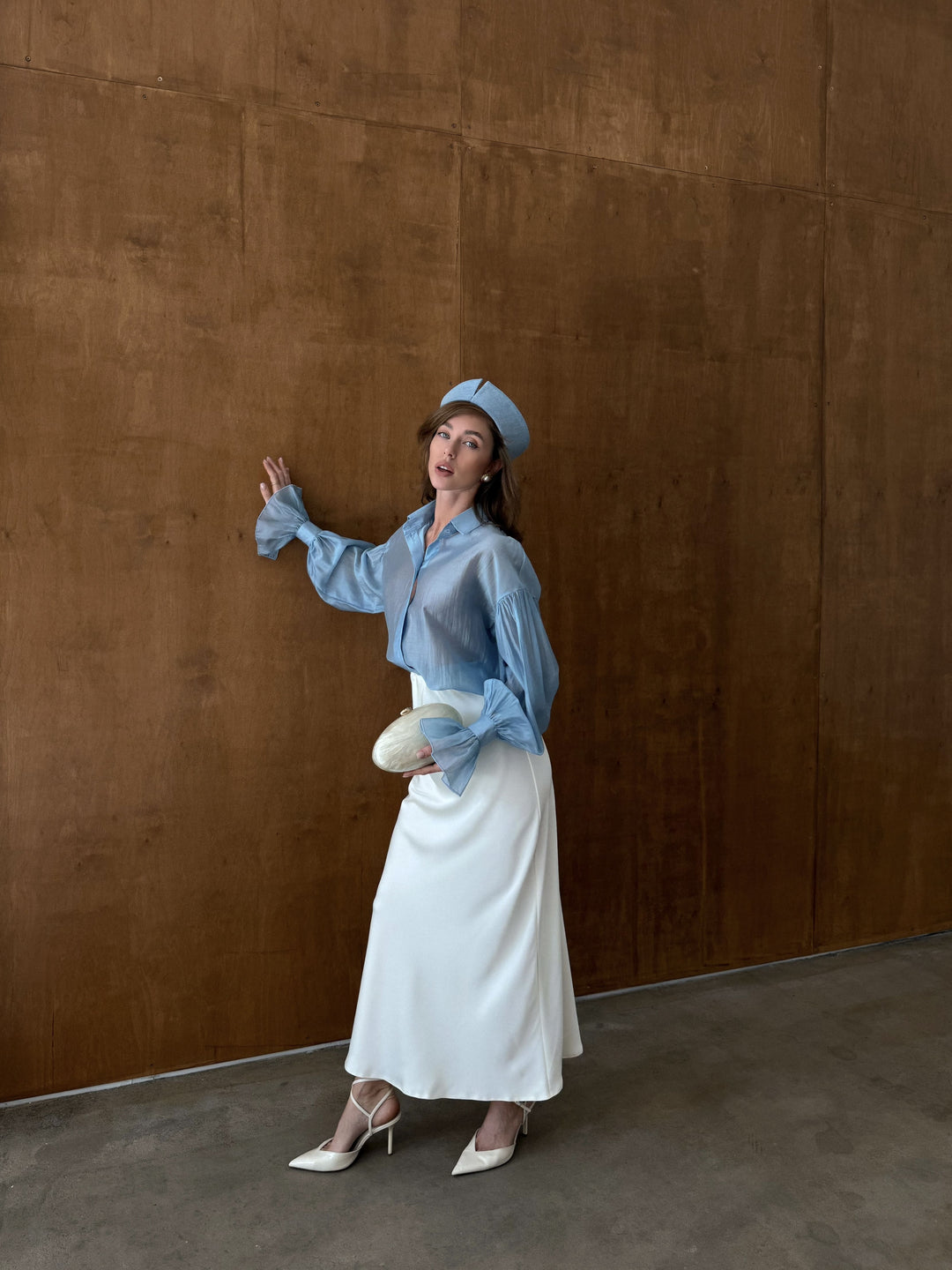 Woman in a blue shirt and ivory satin skirt posing against a brown wall.