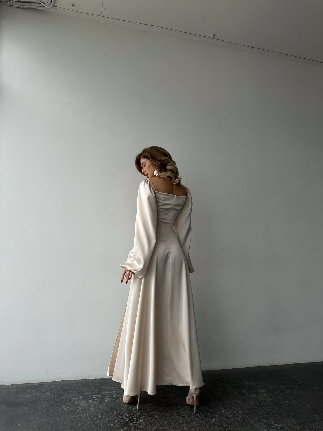 Ivory silk satin maxi dress back view, model looking over shoulder — bishop sleeves, fitted back, floor-length skirt. Perfect for brides, elopements, bridesmaid looks, and formal black-tie events.