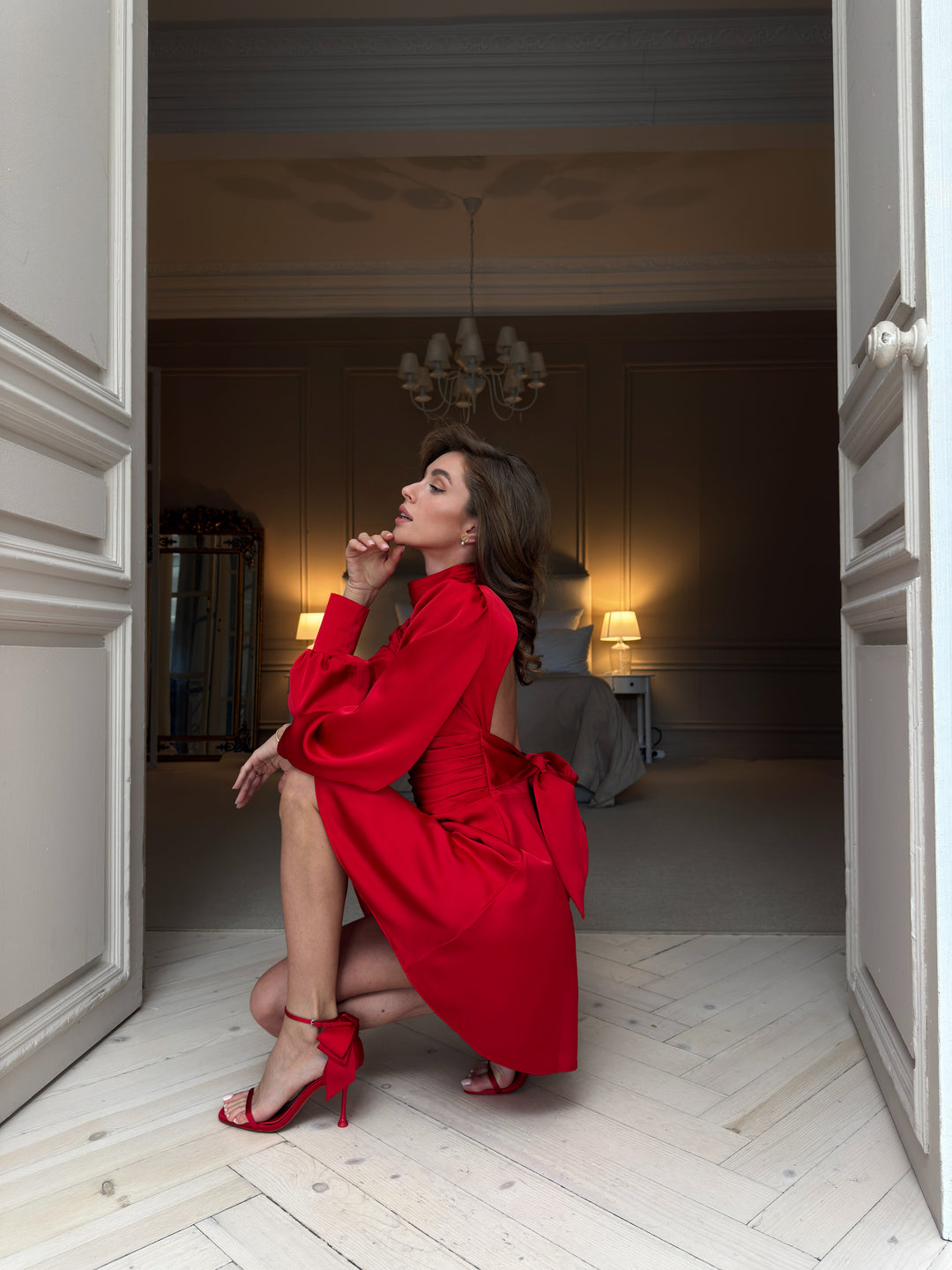 Red silk satin mini dress in profile: pleated high neck, open back, puffed long sleeve, pleated waistband and bow tie falling over the flared skirt.