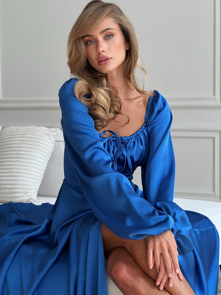 Model posing in a royal blue satin evening gown. The dress features long puff sleeves, a structured corset-style midriff, and a sweeping maxi-length skirt with a thigh slit. An elegant choice for an upscale evening reception.