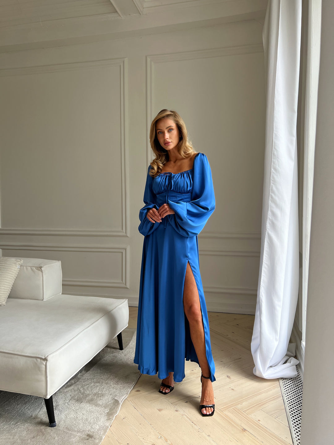 Luxurious long-sleeve royal blue evening gown made of smooth satin. The design includes a flattering ruched bodice, cuffed balloon sleeves, and a sweeping maxi skirt with a side slit. Perfect premium formalwear for a charity ball, prom, or sophisticated cocktail party.