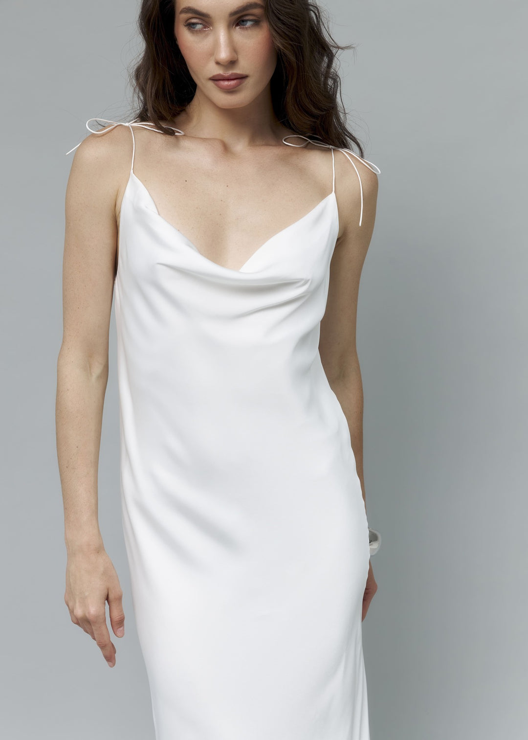 Close-up of white satin bridal slip dress with bow-tie strap and cowl neckline, model placing hand on chest